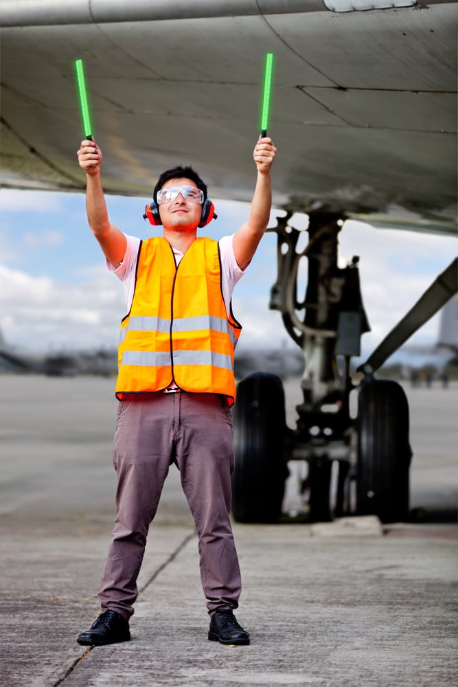 man in orange reflective vest uses light-up sticks to signal incoming planes; the landing gear and bottom of an airplane are visible in the background